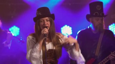 Genç bayan solist, müzik grubuyla sahnede dururken mikrofona şarkı söylüyor. İrlanda tarzı kostümlü rock grubu koyu renk stüdyo ve mavi neon ışıklara karşı. Enerjik müzik. Kapat..