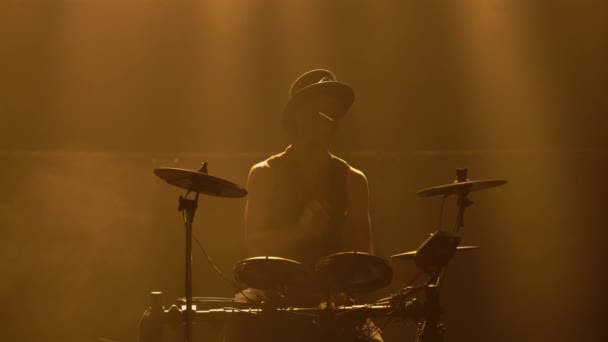 En professionell musiker spelar trummor och slagverk cymbaler i en mörk rökig studio med gula ljus. En trummis i originalkläder och en hatt knackar på ett trumset. Silhuett. Närbild. — Stockvideo