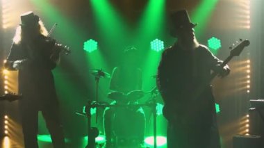 Rock müzisyenleri, koyu renkli dumanlı stüdyoda orijinal İrlanda kostümleri ve şapkalarıyla canlı performans sergiliyorlar. Bir grup erkek elektirik, bas gitar, keman, davul ve sentezleyici çalıyor. Siluetler. Kapat..