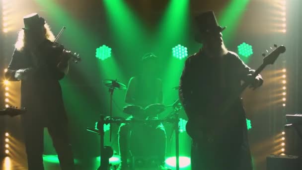 Músicos de rock actúan en vivo con trajes y sombreros irlandeses originales en un oscuro estudio ahumado con luces verdes. Grupo de hombres toca eléctrico, bajo guitarras, violín, batería y sintetizador. Siluetas. De cerca.. — Vídeos de Stock