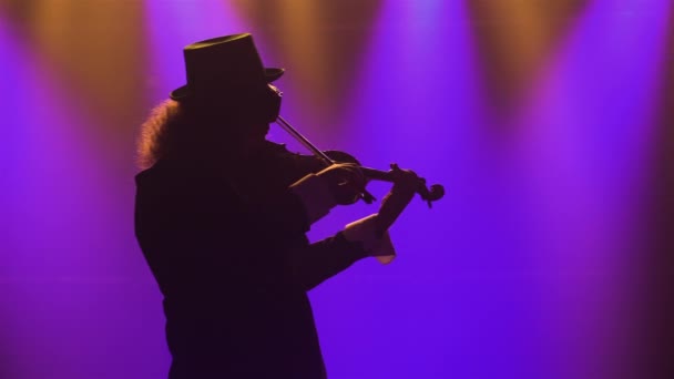 Een man in een retro pak, hoed en originele bril speelt viool. Een muzikant voert een klassiek concert uit in een donkere rokerige studio met blauw licht. Silhouet. Sluiten.. — Stockvideo