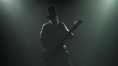 Duman ve neon ışıklarla bir stüdyo gösterisinde sakallı bir rock gitaristi solo bir rol oynar. Şapkalı ve uzun deri pelerinli bir adamın silueti gitar çalıyor. Kapat..
