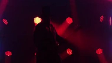 Siyah şapkalı ve pelerinli rock gitaristinin silueti renkli dinamik ışıklarla sahnede performans sergiliyor ve bas gitarda solo performans sergiliyor. Müzisyen ritme ayak uydurmayı sever. Kapat..