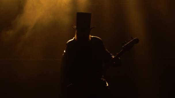 Dynamischer Auftritt eines Rockmusikers, der inmitten von Rauch und leuchtend gelben Neonlichtern Bassgitarre spielt. Mann in Ledermantel und Hut gibt Live-Konzert. Silhouette. Nahaufnahme. — Stockvideo