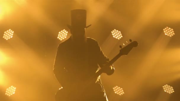 煙と明るい黄色のネオンの中でベースギターを演奏するロックミュージシャンによるダイナミックなパフォーマンス。革のコートと帽子の男はライブコンサートを行います。シルエット。閉じろ!. — ストック動画