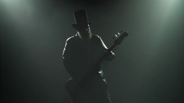Vousatý rockový baskytarista hraje sólovou roli během studiového představení s kouřovými a neonovými světly. Na kytaru hraje silueta muže v klobouku a dlouhém koženém plášti. Zavřít. — Stock video
