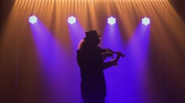 Retro takım elbiseli, şapkalı ve orijinal gözlüklü bir adam keman çalıyor. Bir müzisyen, koyu dumanlı ve mavi ışıklı bir stüdyoda klasik bir konser veriyor. Siluet. Kapat..