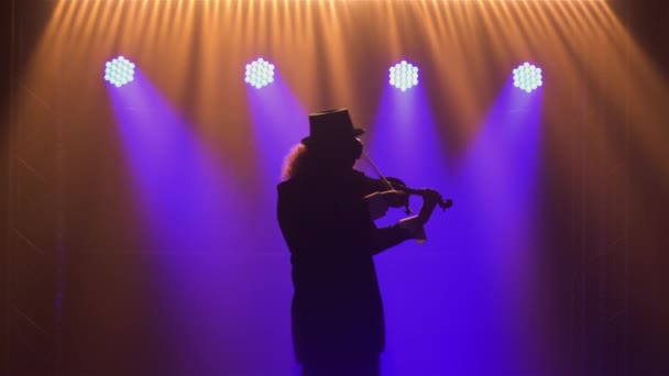Un bărbat în costum retro, pălărie și ochelari originali cântă la vioară. Un muzician susține un concert clasic într-un studio de fum întunecat cu lumini albastre. Silueta. Close up . — Videoclip de stoc