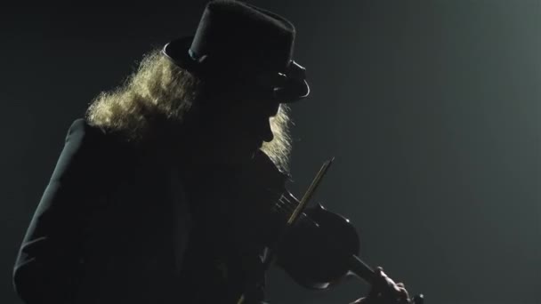 Houslistický hudebník hraje na hudební nástroj. Silueta muže v černém obleku a irském klobouku, hrající melodii na housle. Kouřové tmavé studio s neonovými světly. Zavřít. — Stock video