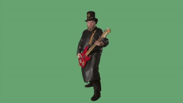 긴 가죽 코트를 입고 아일랜드식 모자를 쓴 수염을 가진 남자가 빨간 기타를 연주 한다. 록 음악가는 녹색 화면 크로마 키로 스튜디오에서 베이스 기타를 연주 한다. 느린 동작. — 비디오