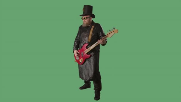 一个留着胡子的男人穿着一件长长的皮革外套，戴着爱尔兰风格的帽子，戴着原始的眼镜，弹奏着红色的吉他。摇滚音乐人在工作室用绿色屏幕彩色键演奏低音吉他。慢动作. — 图库视频影像