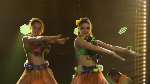 Taneční skupina tančí v krátkých sukních, trikoty zdobené květinami a věnci lei. Okouzlující mladé ženy s pružnými těly pohybují boky na pozadí jasného světla. Zavřít. — Stock video