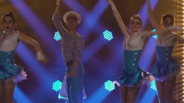 Baştan çıkarıcı takım elbiseli üç güzel kadın denizci şapkalı eşleriyle ateşli bir şekilde dans ediyor. Mavi ışıkların arka planına karşı denizcilik tarzında bir tiyatro gösterisi. Kapat..