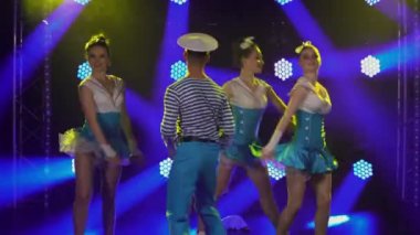 Baştan çıkarıcı takım elbiseli üç güzel kadın denizci şapkalı eşleriyle ateşli bir şekilde dans ediyor. Mavi ışıkların arka planına karşı denizcilik tarzında bir tiyatro gösterisi. Kapat..