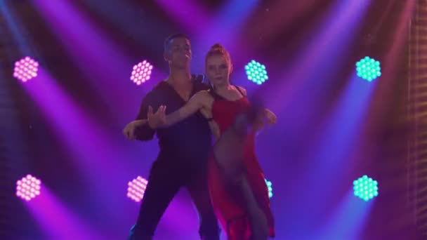 Pasjonująca para tańczących elementów argentyńskiego tanga w ciemnym studio z niebieskimi światłami. Sylwetki mężczyzny w czarnym garniturze i kobiety w czerwonej sukience tańczącej na balu towarzyskim. Zamknij się.. — Wideo stockowe