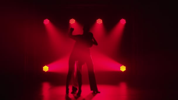 Casal jovem dançando elementos de dança de salão. Rumba, salsa, tango, flamenco. Silhueta de parceiros de dança homem e mulher dançam apaixonadamente no escuro contra o pano de fundo dos holofotes vermelhos. Movimento lento. — Vídeo de Stock