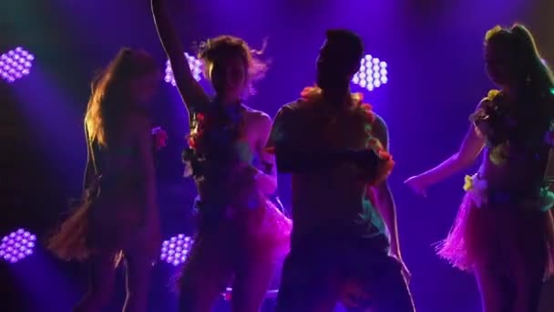 Силуэты трех очаровательных женщин, танцующих вокруг мужчины, сидящего на стуле. Танцевальная группа в гавайском стиле с леями вокруг шеи выступает в тёмной студии с цветными огнями. Закрывай. Медленное движение. — стоковое видео