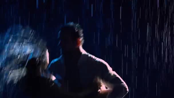 幸せな恋人たちは雨の中で情熱のダンスを実行し、お互いをお楽しみください。水はぬれた服や体を走り、スタジオの光の中で輝きます。幸せなカップルの愛の物語。シルエット。閉じろ!. — ストック動画
