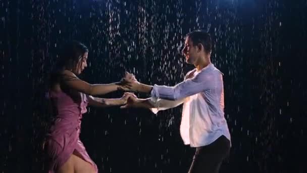Glada älskare utför dans av passion i regn och njuta av varandra. Vatten rinner ner våta kläder och kroppar och gnistrar i studioljus. Kärlekshistoria om lyckliga par. Silhuetter. Närbild. Långsamma rörelser. — Stockvideo