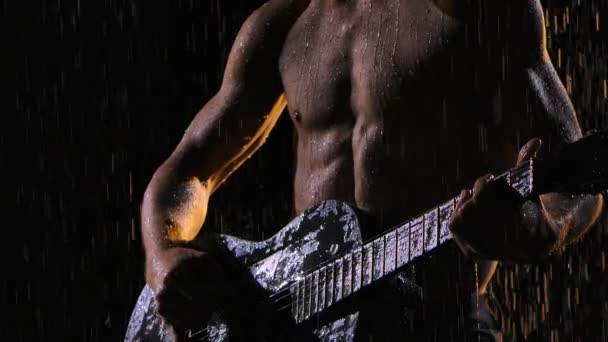 Um homem com um tronco nu, molhado de gotas de chuva, toca guitarra enquanto está sentado na superfície da água. O músico toca violão à chuva, romance, noite. Fecha. Movimento lento. — Vídeo de Stock