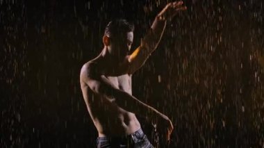 Çıplak gövdeli ıslak bir adam cinsel olarak hareket ediyor, yağmur damlaları kırıyor. Stüdyo ışıklarıyla yağmur derelerinin altında siyah bir zemin üzerinde gitar çalan bir erkeğin silueti. Kapatın. Yavaş çekim.