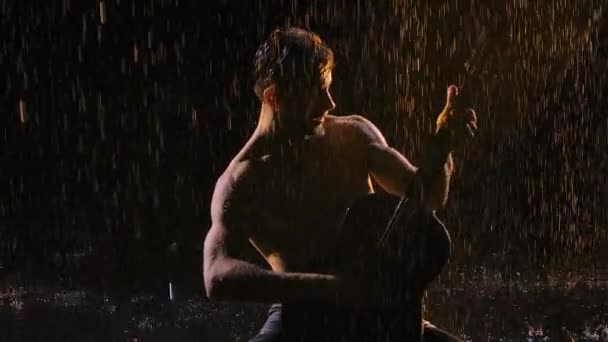 Een man met een naakte romp, nat van regendruppels, speelt gitaar terwijl hij op het wateroppervlak zit. De muzikant speelt gitaar in de regen, romantiek, nacht. Sluit maar af. Langzame beweging. — Stockvideo