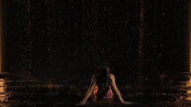 Islak bir elbise içinde poz veren güzel, seksi esmer ve bir tutam saç üretiyor. Dişi, karanlık bir odada stüdyo ışığının önünde yağmurda poz verir ve bundan zevk alır. Yavaş çekim.