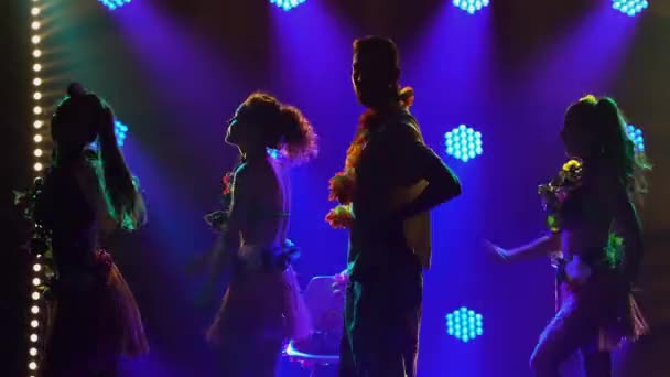 Sylwetki trzech uroczych kobiet tańczących wokół mężczyzny siedzącego na krześle. Grupa taneczna w stylu hawajskim z lei wokół szyi występują w ciemnym studio z kolorowymi światłami. Zamknij się.. — Wideo stockowe