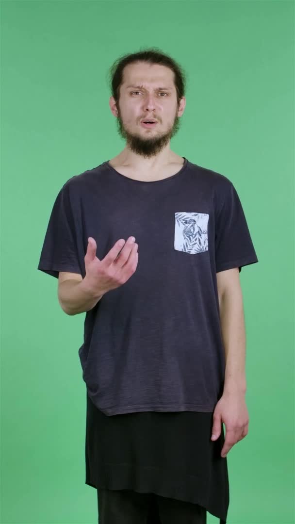 Portrait d'un homme regardant la caméra, agitant les mains dans l'indignation, comment est-ce. Un homme brun avec une barbe dans un T-shirt noir dans le studio sur un écran vert. Au ralenti. Vidéo verticale. — Video