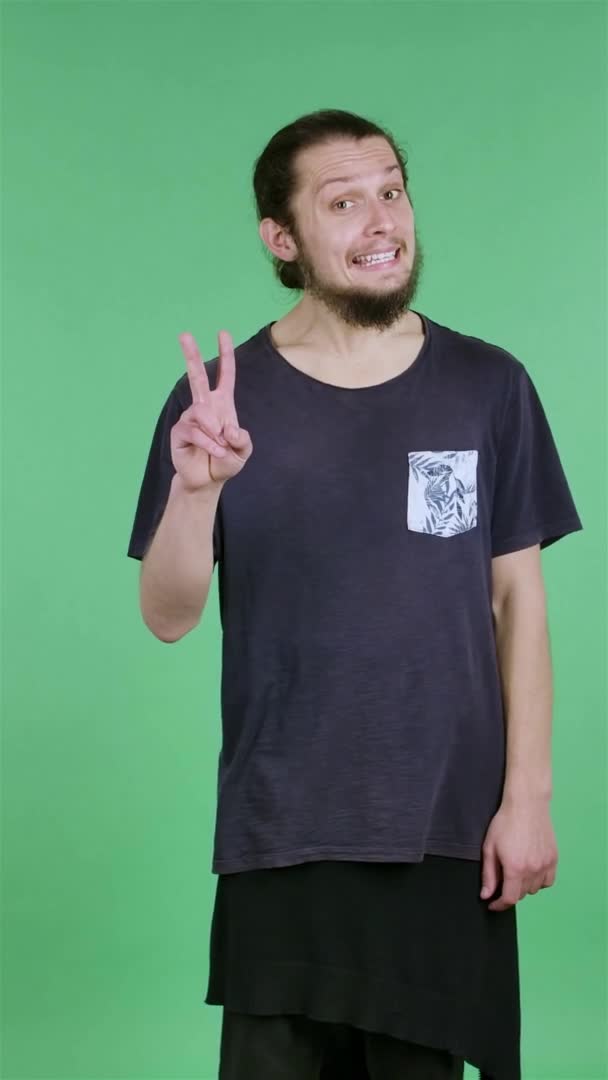 Le portrait d'un homme montre un geste de victoire avec deux doigts. Un geste d'approbation, de paix, de convivialité, de salutation. Homme barbu aux cheveux foncés dans le studio sur un écran vert. Au ralenti. Vidéo verticale. — Video