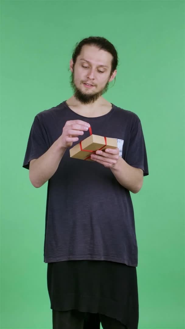 Langsam öffnet ein Mann die Geschenkbox, faltet die Geburtstagsüberraschung auf und drückt sein Erstaunen über das Geschenk aus. Der Typ im Studio auf einer grünen Leinwand. Zeitlupe. Vertikales Video. — Stockvideo