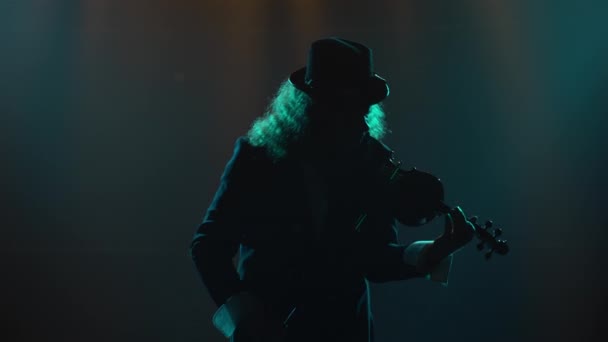 Músico violinista toca un instrumento musical. Silueta de un hombre con traje negro y sombrero irlandés, tocando una melodía en un violín. Estudio oscuro ahumado con luces de neón. De cerca.. — Vídeos de Stock