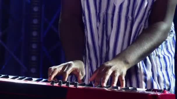 Homem afro-americano toca sintetizador de piano e canta em estúdio escuro com luzes azuis. Músico com padrão étnico branco em seu rosto em camisa listrada nacional se apresenta no palco. Fechar. — Vídeo de Stock