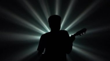 Afrika kökenli Amerikalı bir adamın siyah siluetinin alt görüntüsü, elektro gitar çalıyor. Müzisyen, bir enstrümanın tellerini karanlıkta, dönen parlak ışık demetine karşı çalar. Kapat..