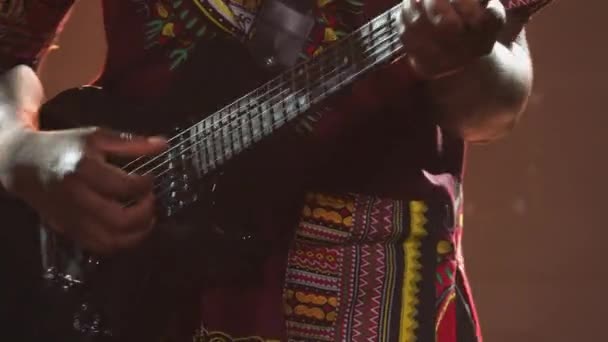 Афроамериканский музыкант в этническом национальном костюме играет на электрогитаре в темной студии. Черный человек закрыл глаза и наслаждается игрой на музыкальном инструменте на фоне огней. Закрыть. — стоковое видео