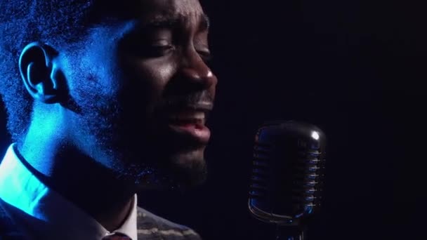 African American man in a stylish suit sings a song into a retro microphone. The jazz vocalist performs in a dark studio with bright blue lights. African ethnic music. Side view. Close up. — Stock Video