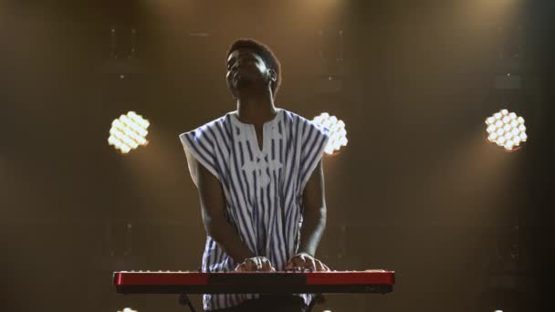 Hombre afroamericano toca el teclado sintetizador de piano y canta en estudio oscuro con luces brillantes. Músico con patrón étnico blanco en su cara en camisa nacional a rayas realiza en el escenario. — Vídeo de stock