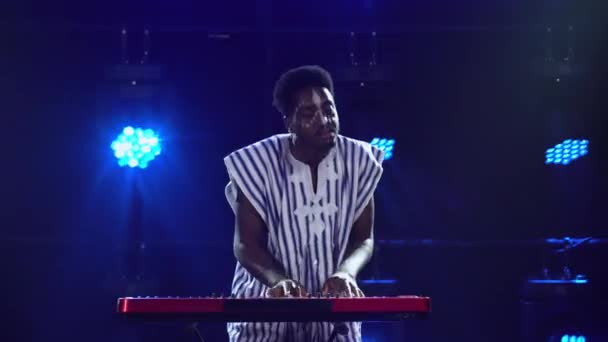 Afro-Américain joue du piano synthétiseur clavier et chante en studio sombre avec des lumières bleues. Musicien avec un motif ethnique blanc sur le visage en chemise à rayures nationales se produit sur scène. — Video