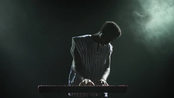 Dark Smoky Stüdyoda sahnede piyano sentezleyicisi çalan Afrikalı Amerikalı adamın silueti. Yüzünde beyaz etnik bir desen olan siyah müzisyen ışıkların arka planına karşı gösteri yapıyor.. — Stok video