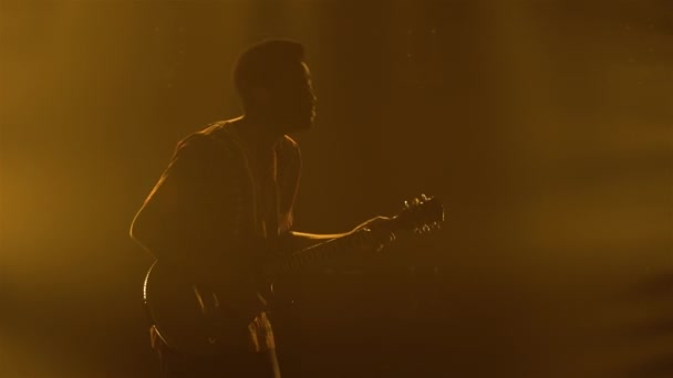 La silhouette dell'uomo afroamericano in abito etnico nazionale suona la chitarra elettrica e balla a ritmo di musica. Chitarrista nero maschio suona uno strumento elettrico in studio di fumo scuro con luci gialle — Video Stock