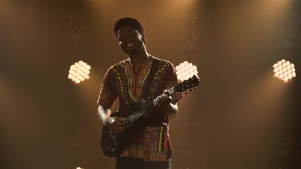 Musicien afro-américain en costume ethnique national joue de la guitare électrique en studio sombre. Homme noir a fermé les yeux et aime jouer de l'instrument de musique sur fond de lumières. — Video