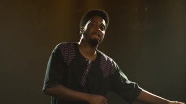 Ulusal etnik kostümlü Afrikalı Amerikalı genç adam zevkle dans ediyor ve bir şarkı söylüyor. Afrika folkloru, siyah bir sanatçı tarafından karanlık bir stüdyoda duman ve sarı ışıkla seslendirildi. Kapat..
