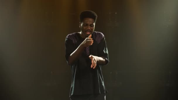 Le jeune homme afro-américain en costume ethnique national danse avec plaisir et chante une chanson. Folklorique africain interprété par un artiste noir dans un studio sombre avec fumée et lumière jaune. — Video