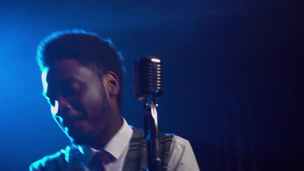 African American man in a stylish suit sings a song into a retro microphone. The jazz vocalist performs in a dark studio with bright blue lights. African ethnic music. Close up. — Stock Video