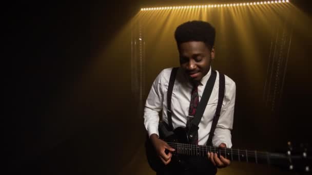 Happy smiling African American young man in stylish suit plays electric guitar and sings a song. The male performs a salty concert in a dark studio against the backdrop of smoke and lights. — Stock Video