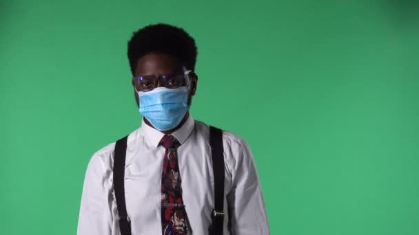 医療用マスクの若いアフリカ系アメリカ人の男性の肖像画は、カメラを見て、マスクを削除し、安心してため息をつく。スタジオの緑のスクリーンにポーズをとっている白いシャツに眼鏡をかけた黒人男性。閉じろ!. — ストック動画