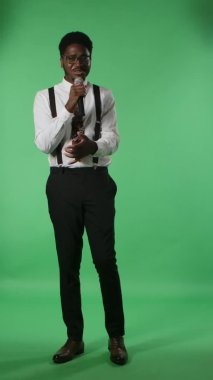 Genç Afrikalı Amerikalı bir adamın portresi mikrofona mutlu bir şekilde şarkı söylüyor ve elleriyle hareket ediyor. Stüdyoda yeşil ekranda poz veren kravatlı ve gözlüklü siyah bir erkek. Dikey video.