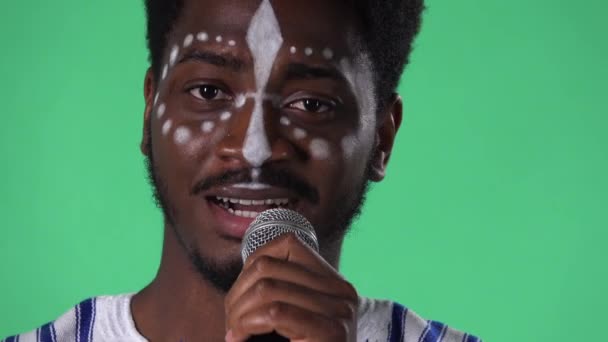 Retrato del joven afroamericano cantando canción en el micrófono. Hombre negro con un patrón étnico blanco en la cara posando en pantalla verde en el estudio. De cerca.. — Vídeos de Stock