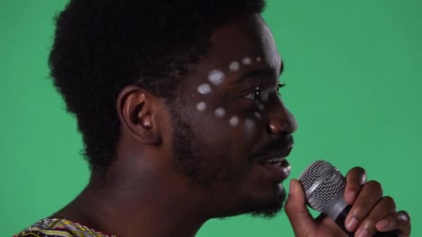 Портрет молодого афроамериканца, поющего песню в микрофон. Чёрный мужчина с этнически белым рисунком на лице позирует на зелёном экране в студии. Вид сбоку. Закрыть. — стоковое видео
