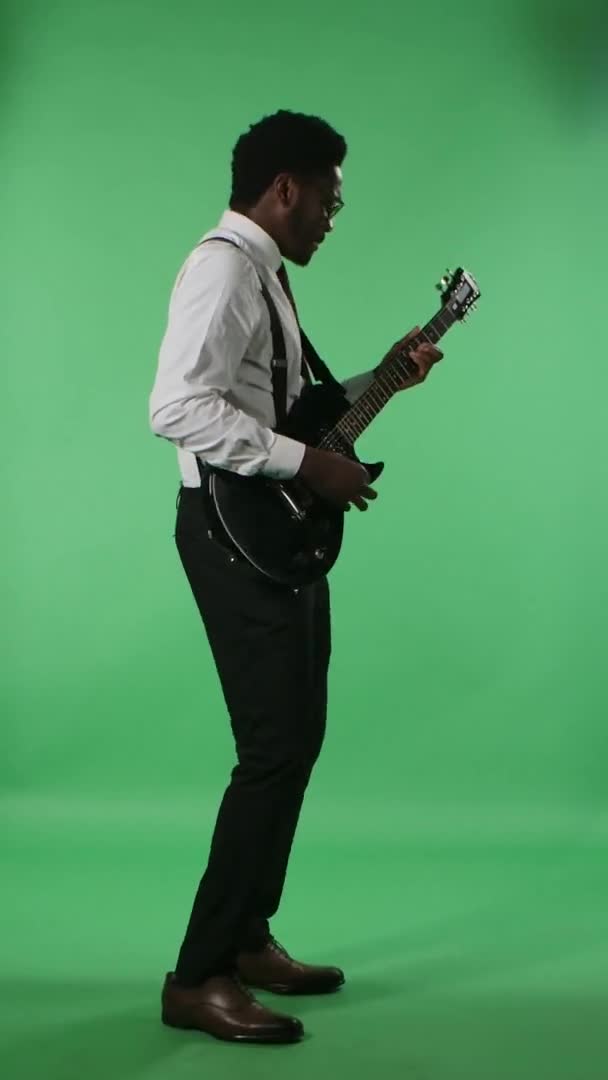 Gülümseyen ve elektro gitar çalan Afro-Amerikan bir adamın portresi. Stüdyoda yeşil ekranda poz veren kravatlı ve gözlüklü siyah bir erkek. Yan görüş. Dikey video. — Stok video